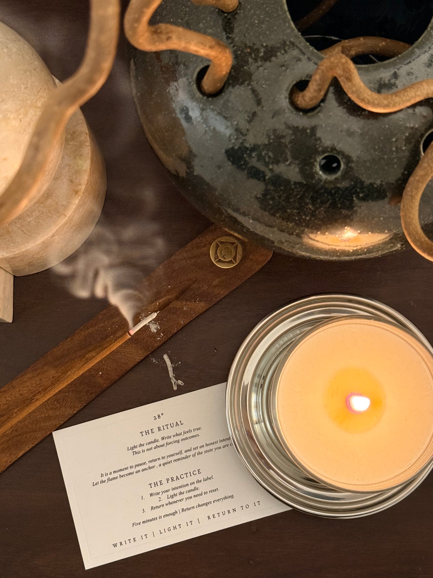 Candle lit on a metal holder with a piece of paper next to it on a reflective surface.