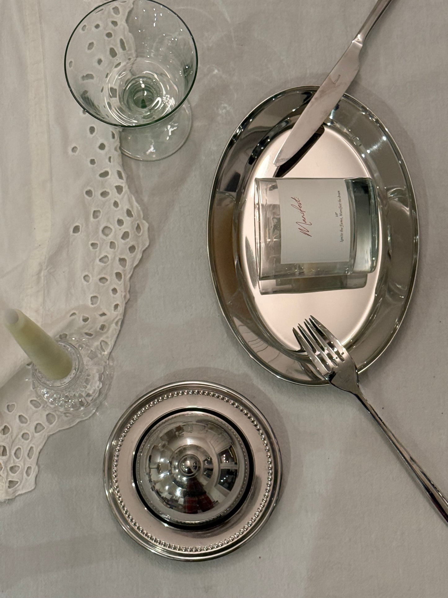 Silver tableware including plates, knife, fork, and glass on a white tablecloth with a  candle on a silver plate.