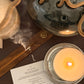 Candle lit on a metal holder with a piece of paper next to it on a reflective surface.